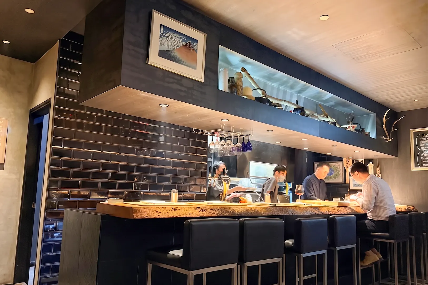 A modern bar with a wooden counter and black stools, featuring a dark tiled wall and an overhead display.