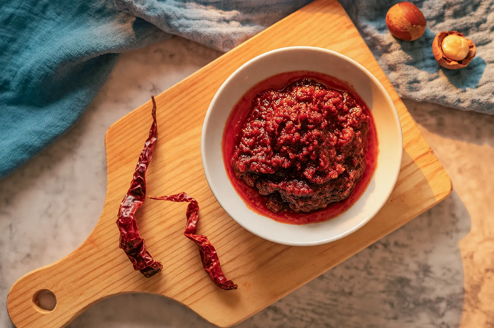 A bowl of red chili paste on a wooden cutting board, with dried chilies beside it. A blue cloth and hazelnuts add warmth and texture to the scene.