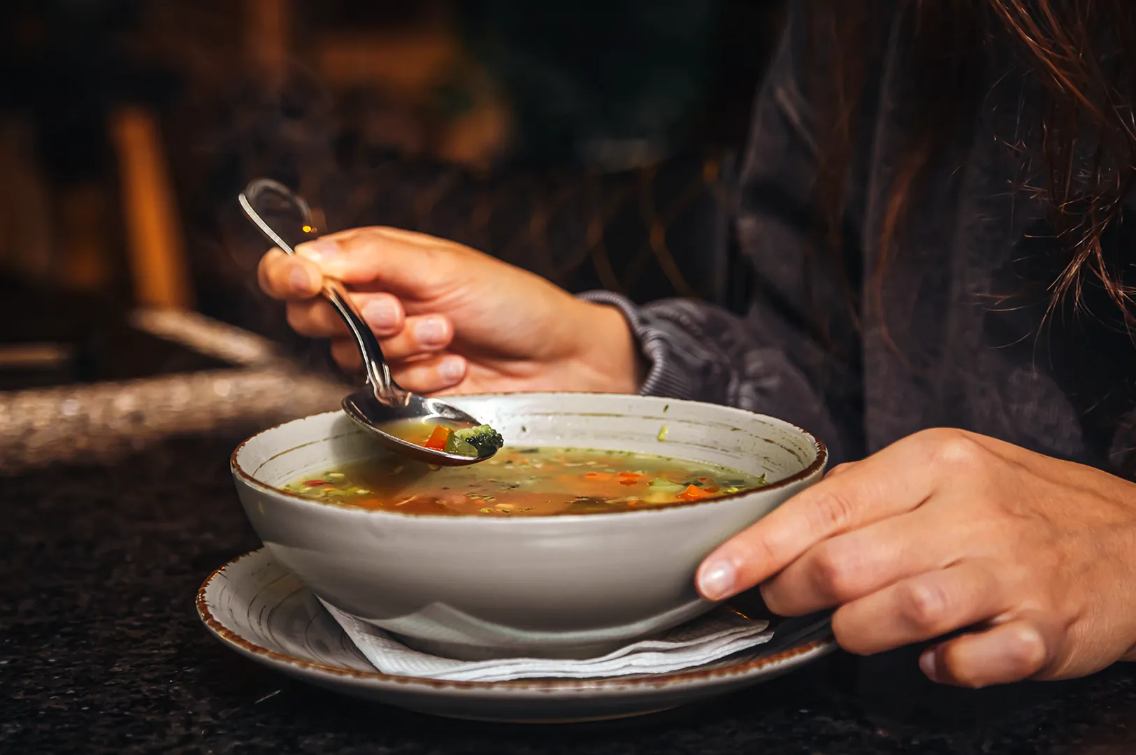 A person holds a spoon above a bowl of vegetable soup, ready to eat. The warm lighting and cozy setting suggest a comforting meal