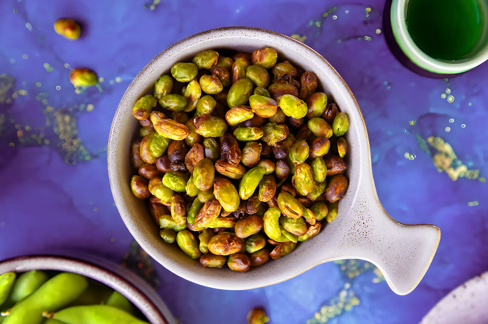 A bowl filled with roasted edamame on a textured blue surface. The edamame are golden and vibrant, with a rustic bowl, creating a fresh and appetizing look.