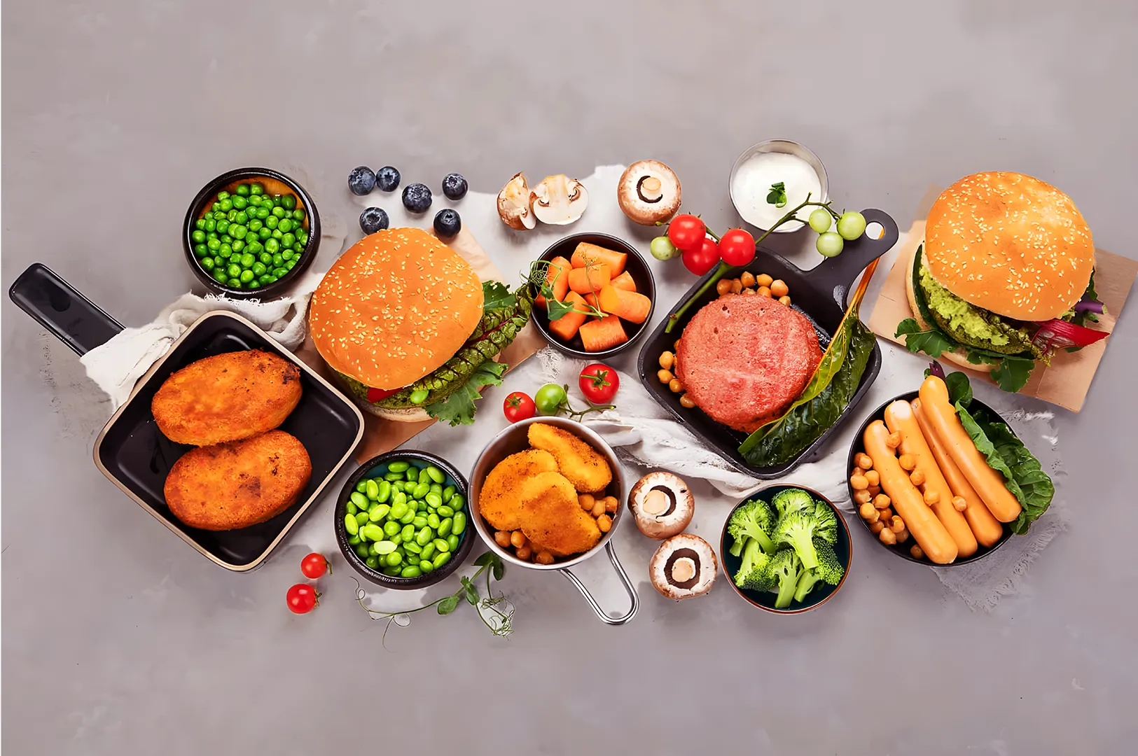 A variety of plant-based foods on a gray background, including burgers, nuggets, sausages, fresh vegetables, and fruits, showcasing a vibrant, healthy display.