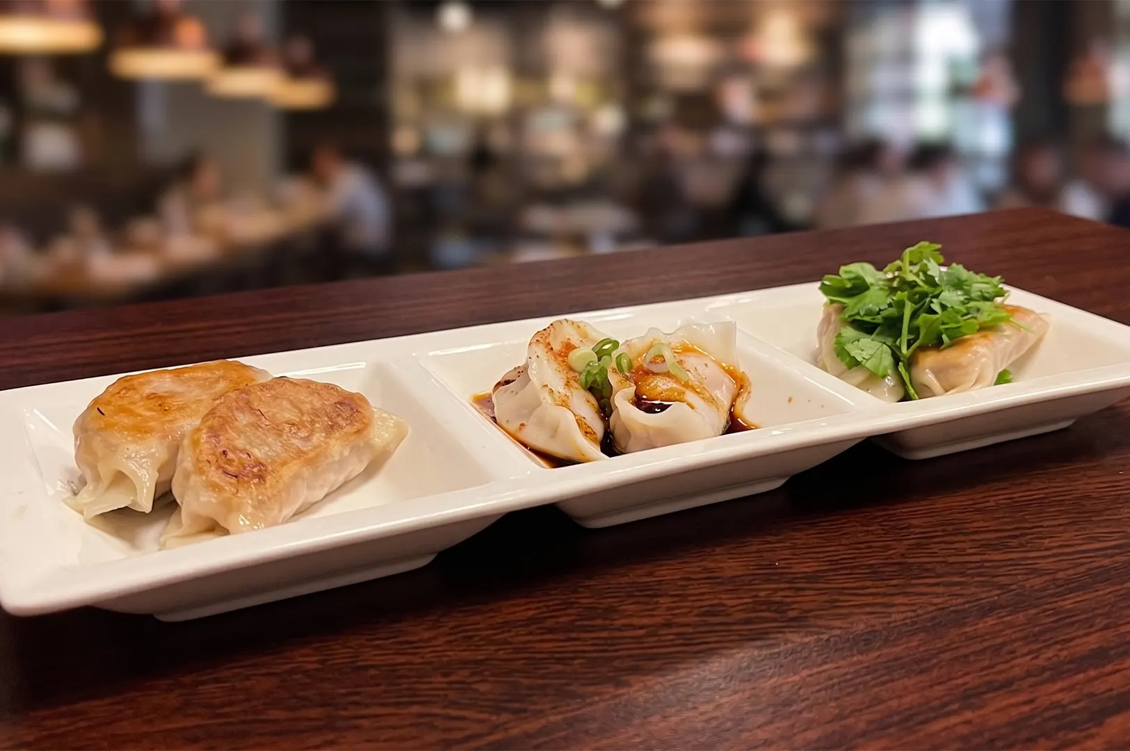 Three varieties of pan-fried dumplings served on a white divided plate in a restaurant setting.