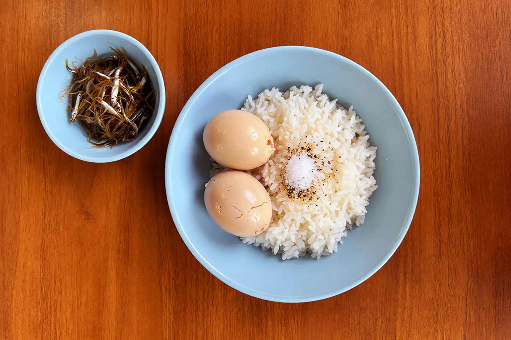 A blue bowl filled with steamed rice topped with two boiled eggs, salt, and pepper sits on a wooden table. A smaller bowl with dried fish is beside it.