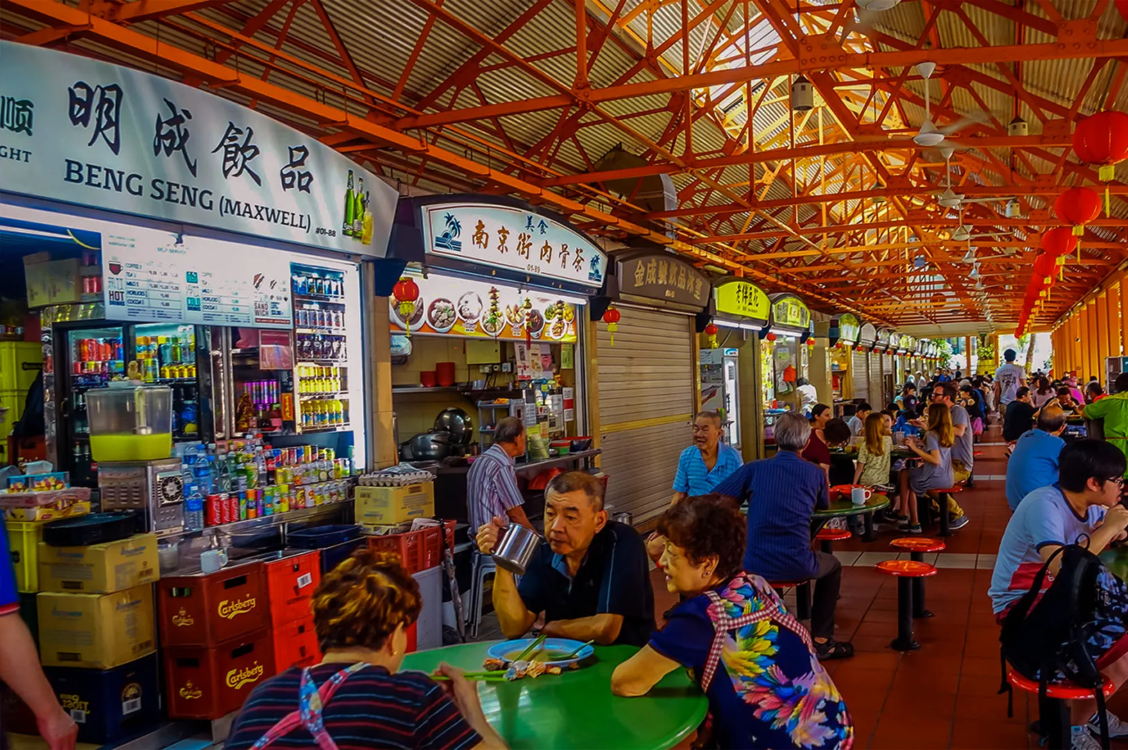 Bustling hawker center with people dining at colorful tables under an orange lattice roof. Food stalls line the background, creating a lively atmosphere