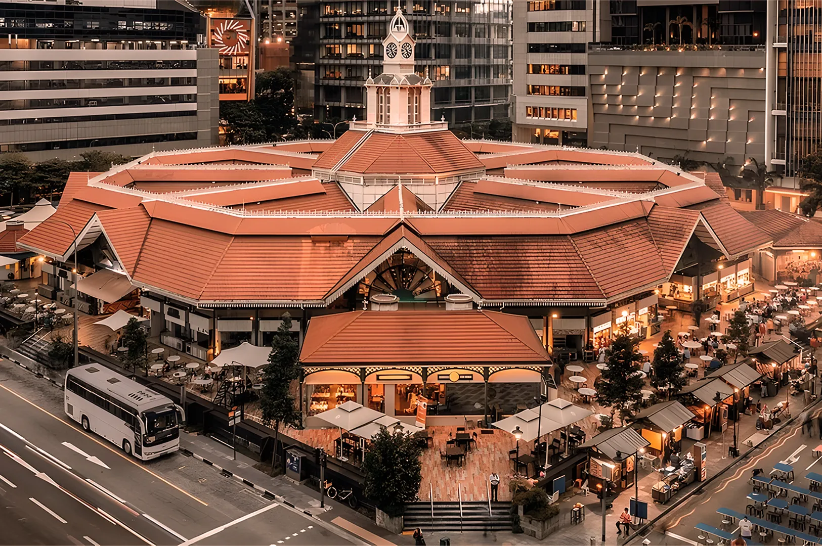 Historic rotunda with red-tiled roof, surrounded by modern buildings. Bustling evening market atmosphere with stalls, people dining, and a bus on the street.