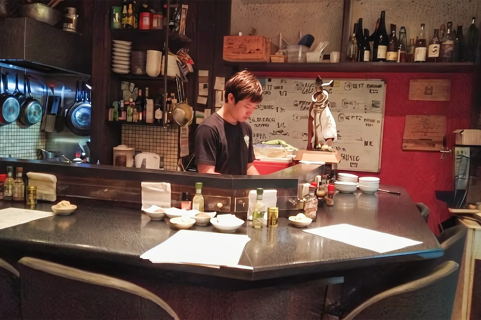 A man is working behind a dark L-shaped bar in a small, rustic restaurant with shelves of bottles and a whiteboard in the background.