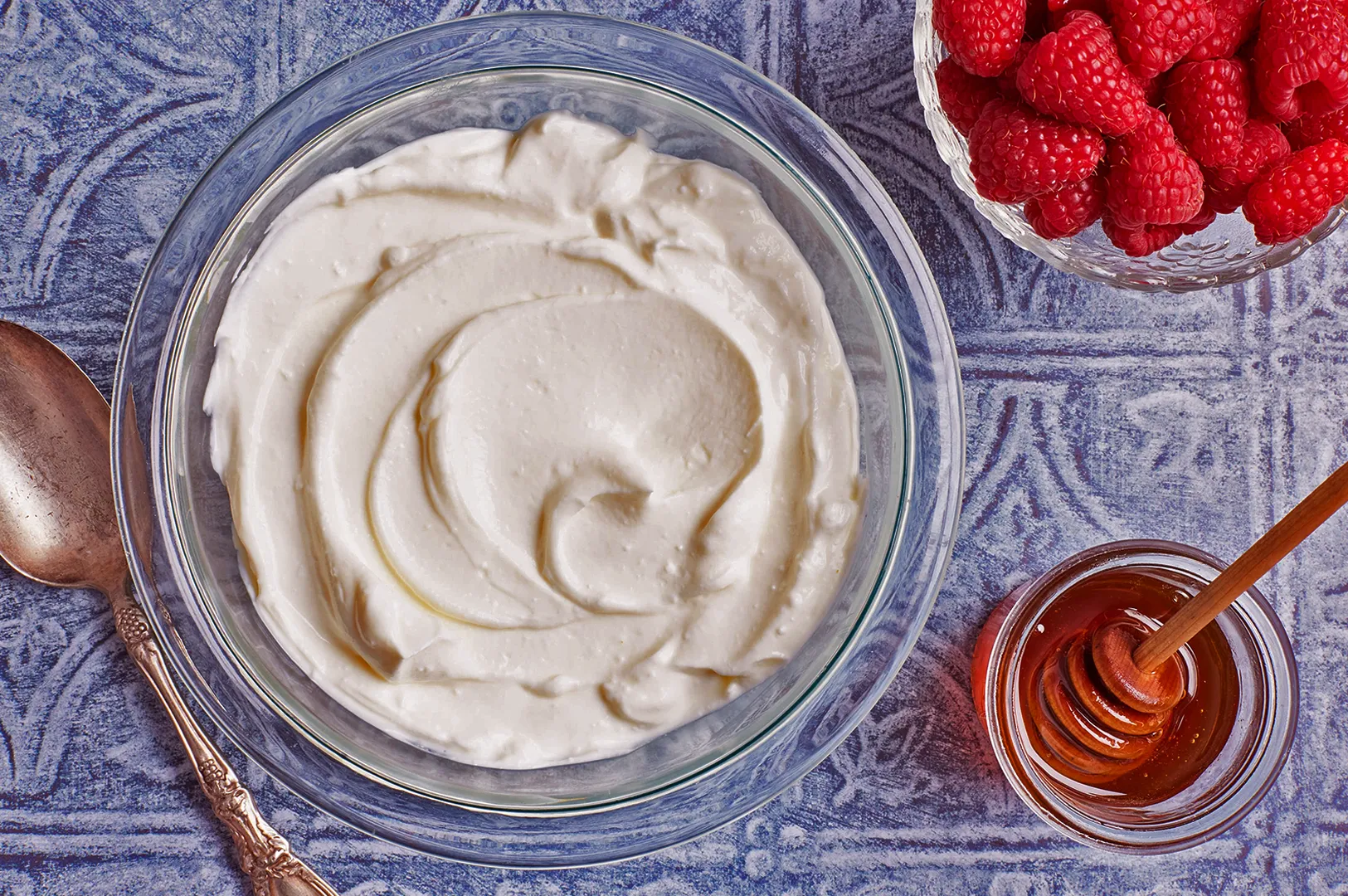 A clear bowl of creamy Greek yogurt is centered on a blue-patterned tablecloth. To its right, bright red raspberries and a small jar of golden honey with a dipper exude freshness and sweetness. A vintage silver spoon lies to the left.
