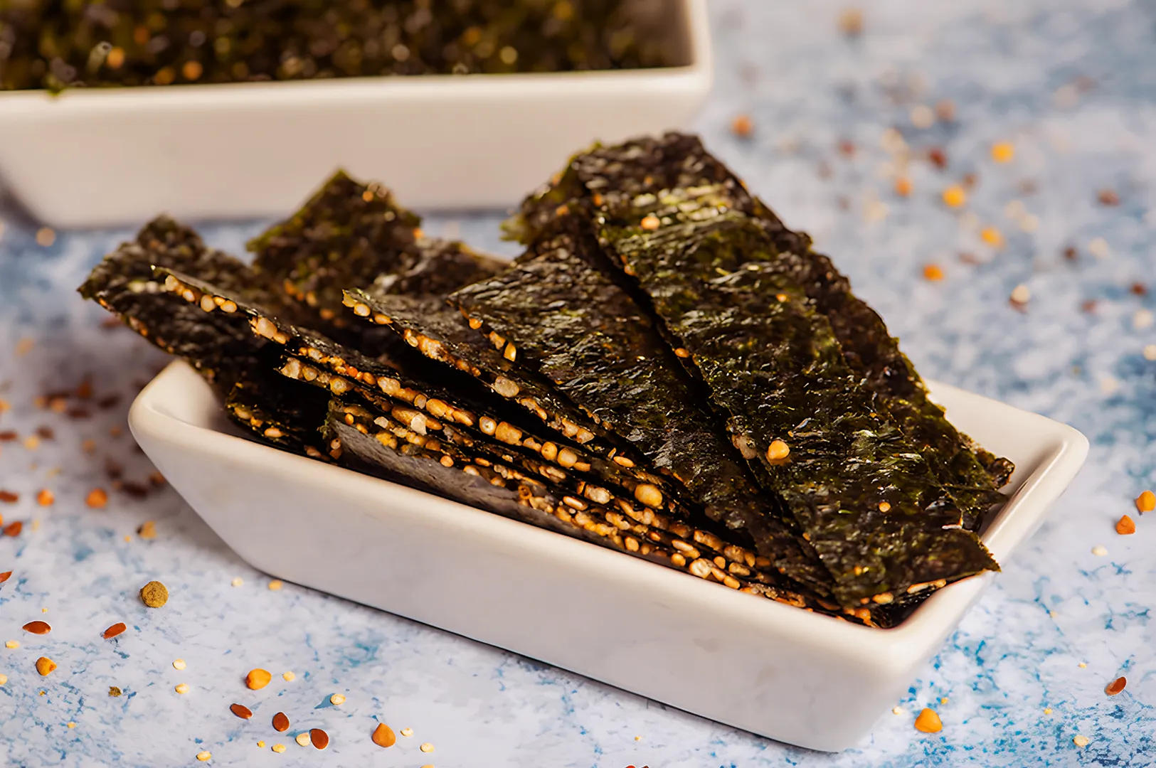 Rectangular seaweed snacks with sesame seeds in a white dish on a speckled blue surface, creating a fresh and appetizing presentation.