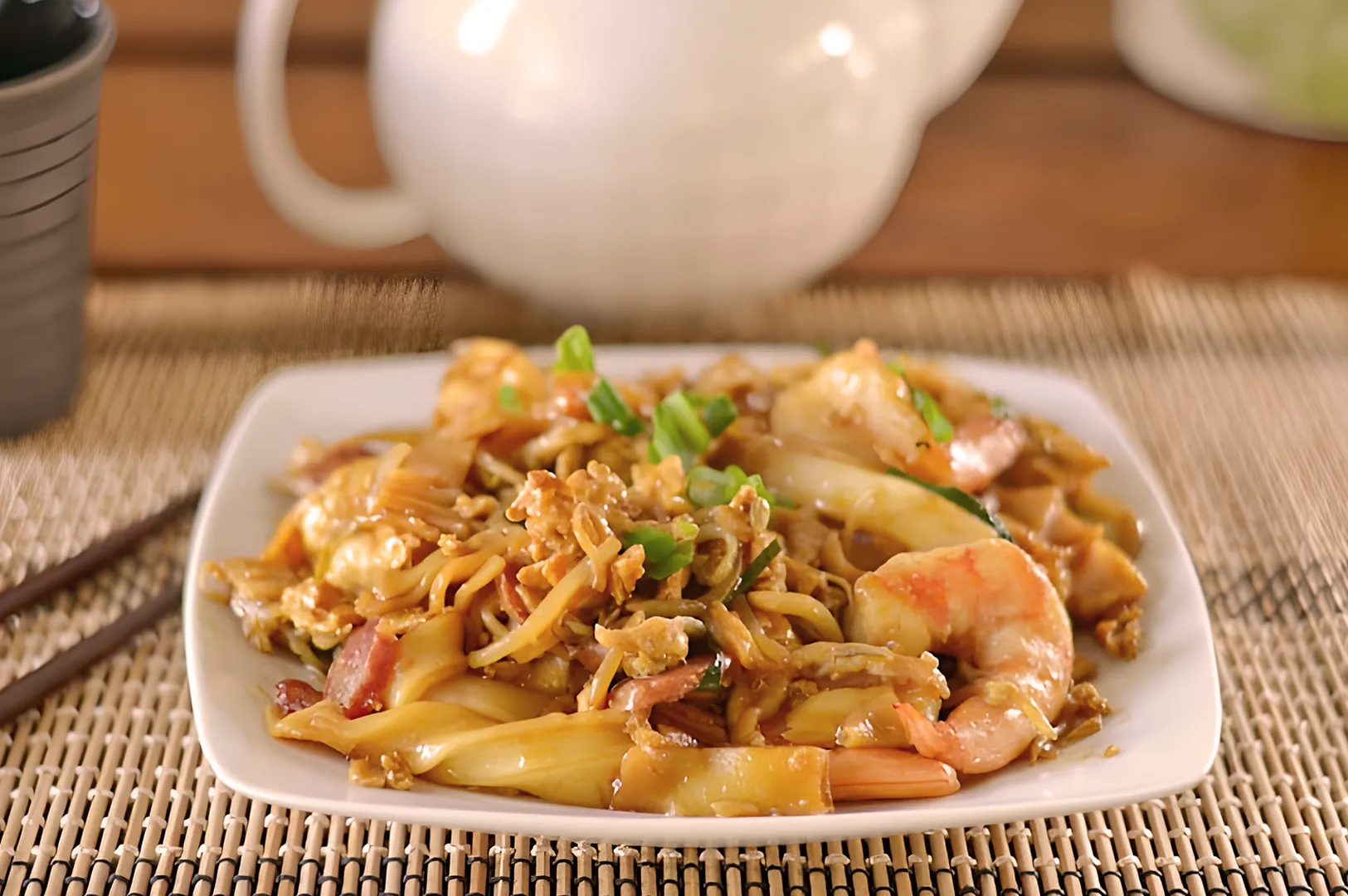 A plate of stir-fried noodles with shrimp, green onions, and crispy bits of egg on a bamboo mat. A teapot and chopsticks are in the background.