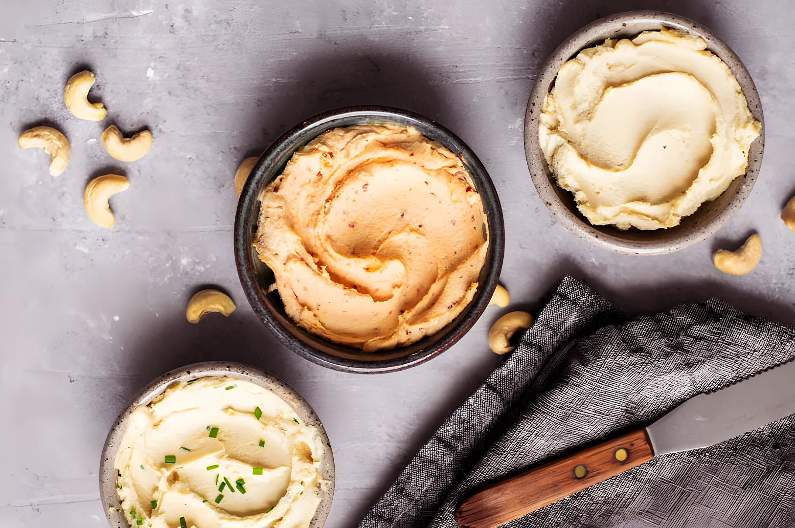 Three bowls of creamy cashew cheese sit on a gray surface. One is plain, one has herbs, and another is spiced. Cashews and a knife are nearby.
