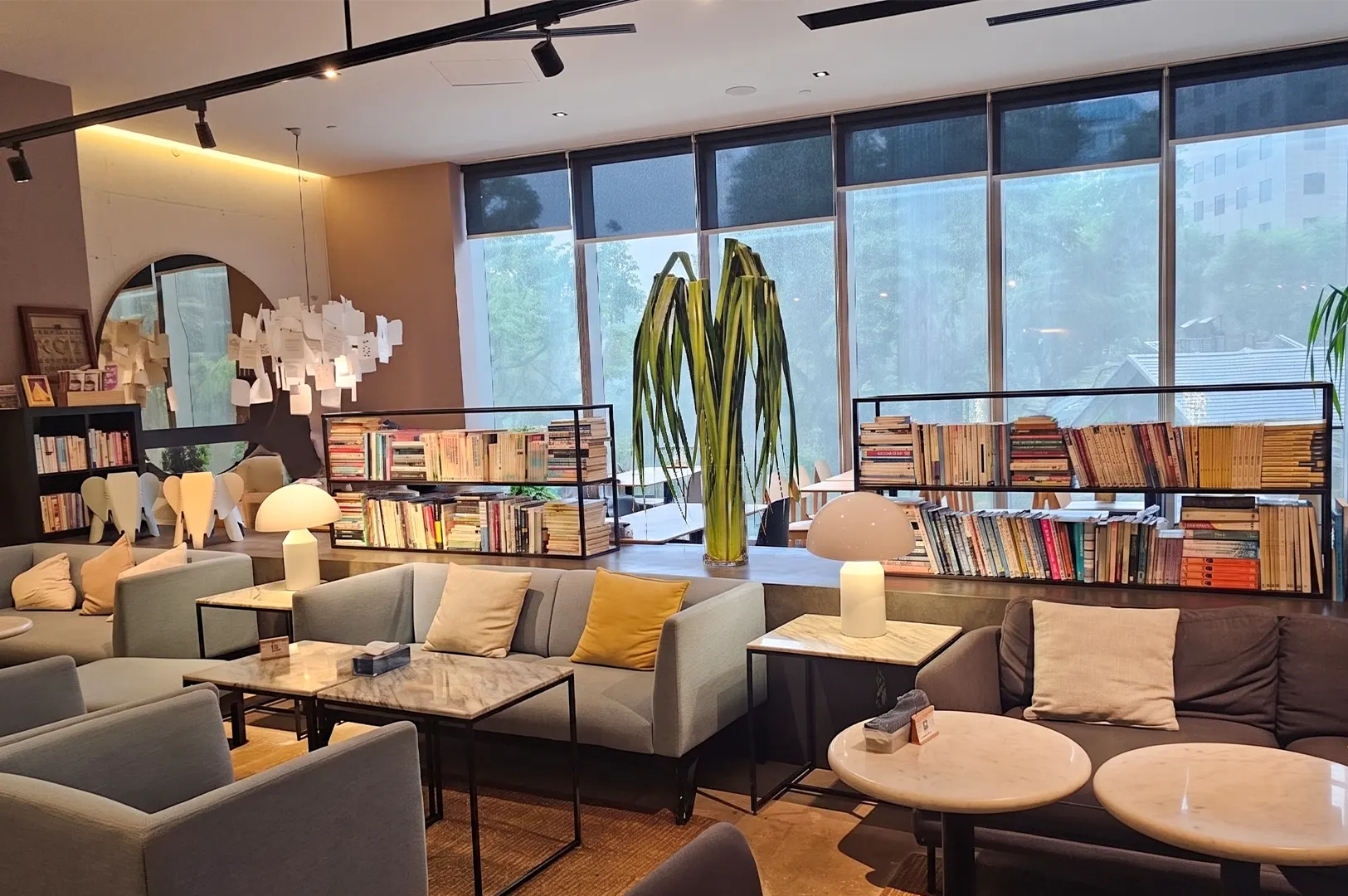A modern lounge area featuring soft blue and grey sofas with marble-top tables, set against a backdrop of bookshelves and large windows.