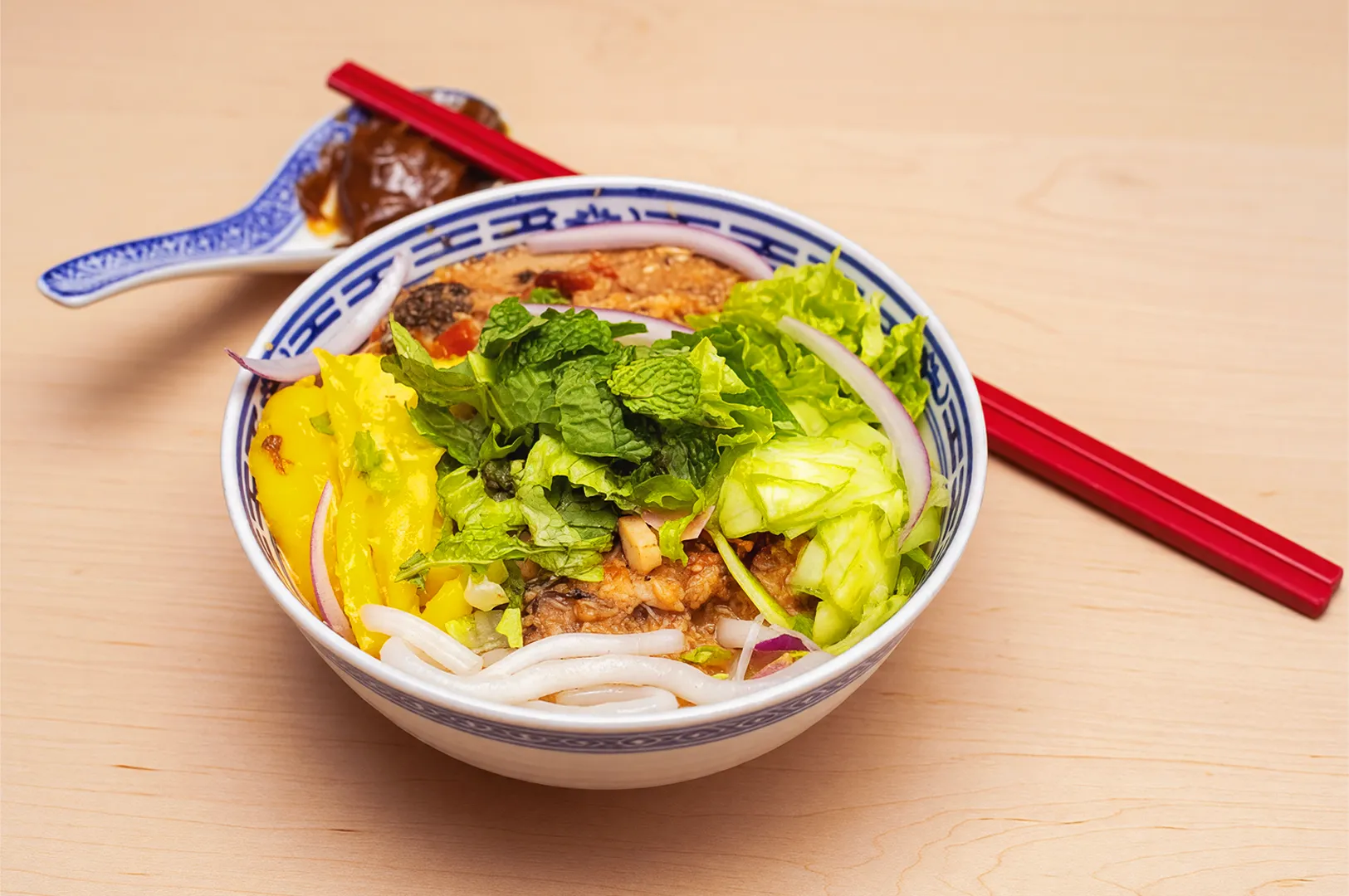 A vibrant bowl of Vietnamese noodle soup with fresh mint, lettuce, red onion, and fish, served with red chopsticks and a sauce on a blue spoon.