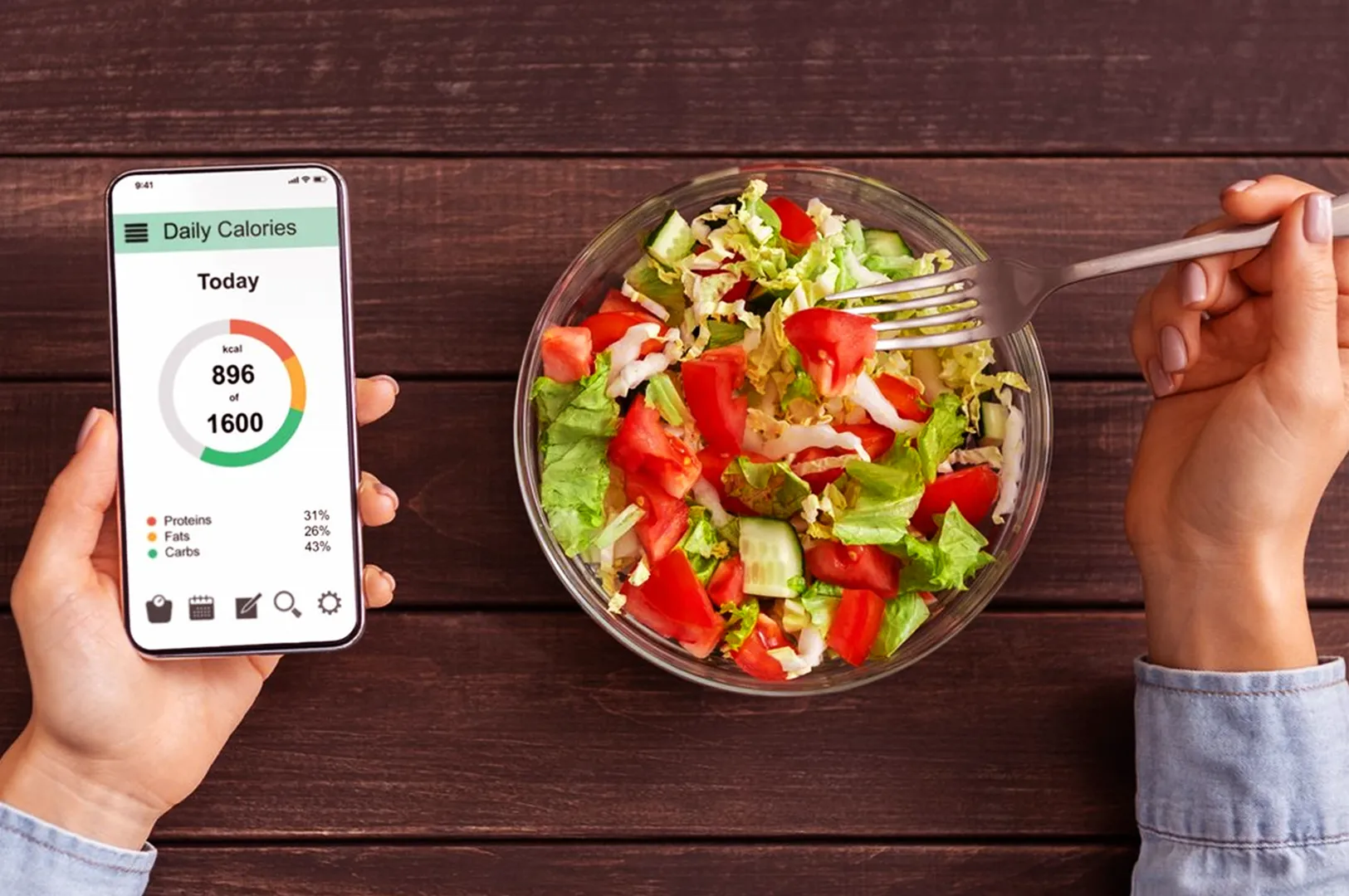 A person holding a smartphone displaying a daily calorie tracker app beside a bowl of fresh salad. The setting conveys a healthy lifestyle focus.
