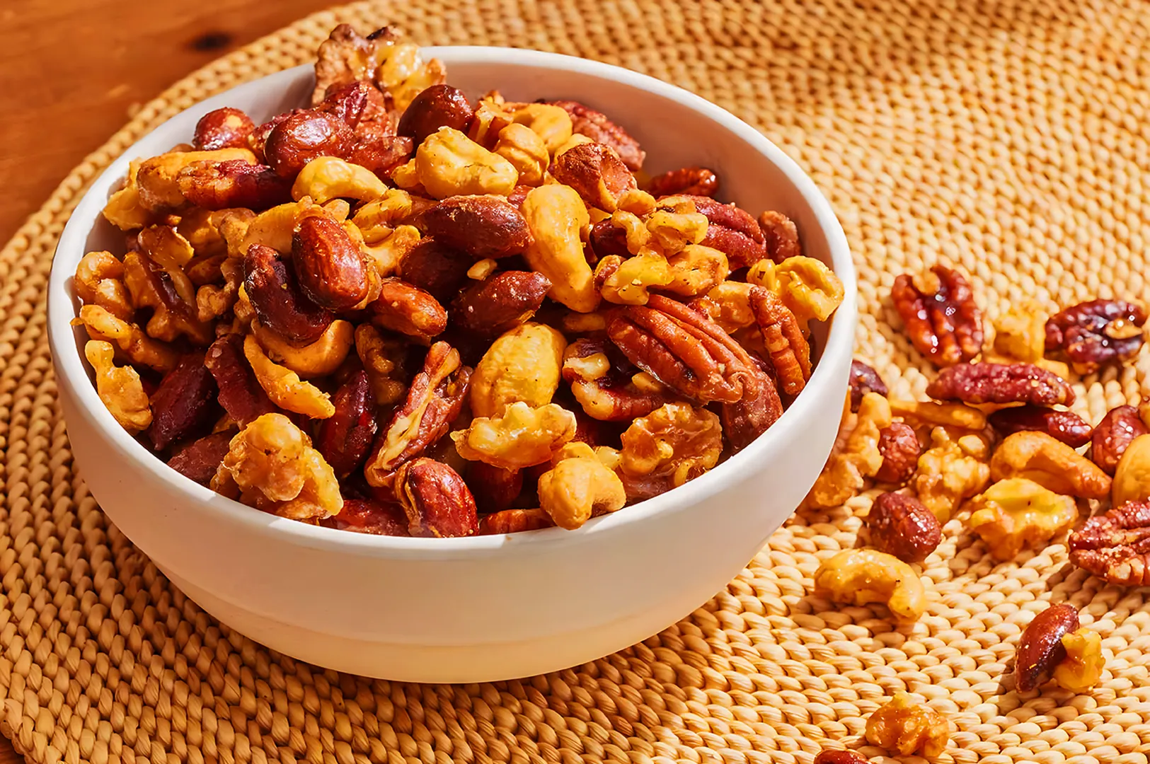 A white bowl filled with assorted nuts, including cashews, almonds, and pecans, sits on a woven mat. Some nuts are scattered around, creating a warm, inviting atmosphere.