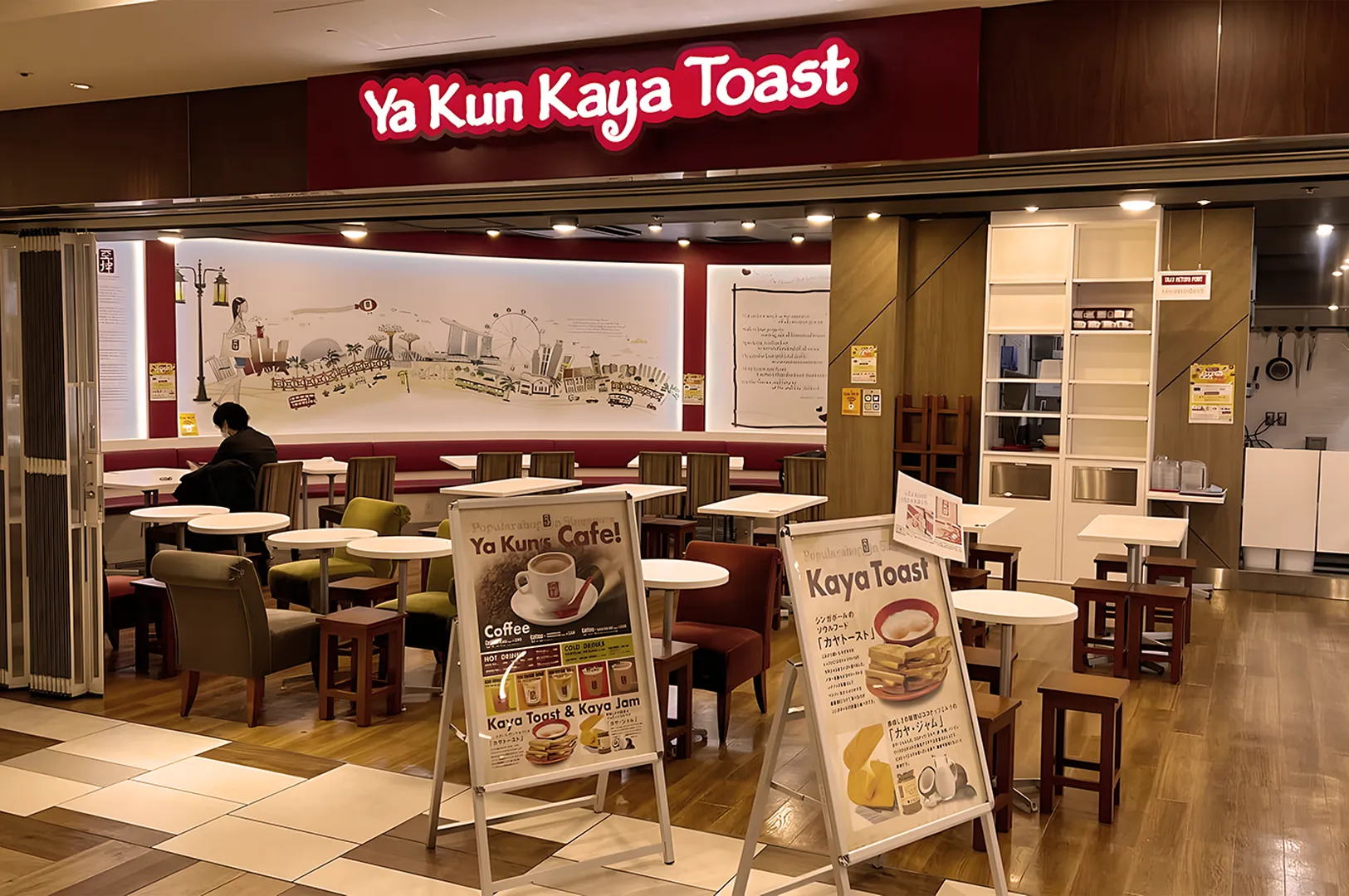 Ya Kun Kaya Toast cafe interior with cozy seating, white tables, and menu boards. A mural adorns the back wall. Warm, inviting ambiance.