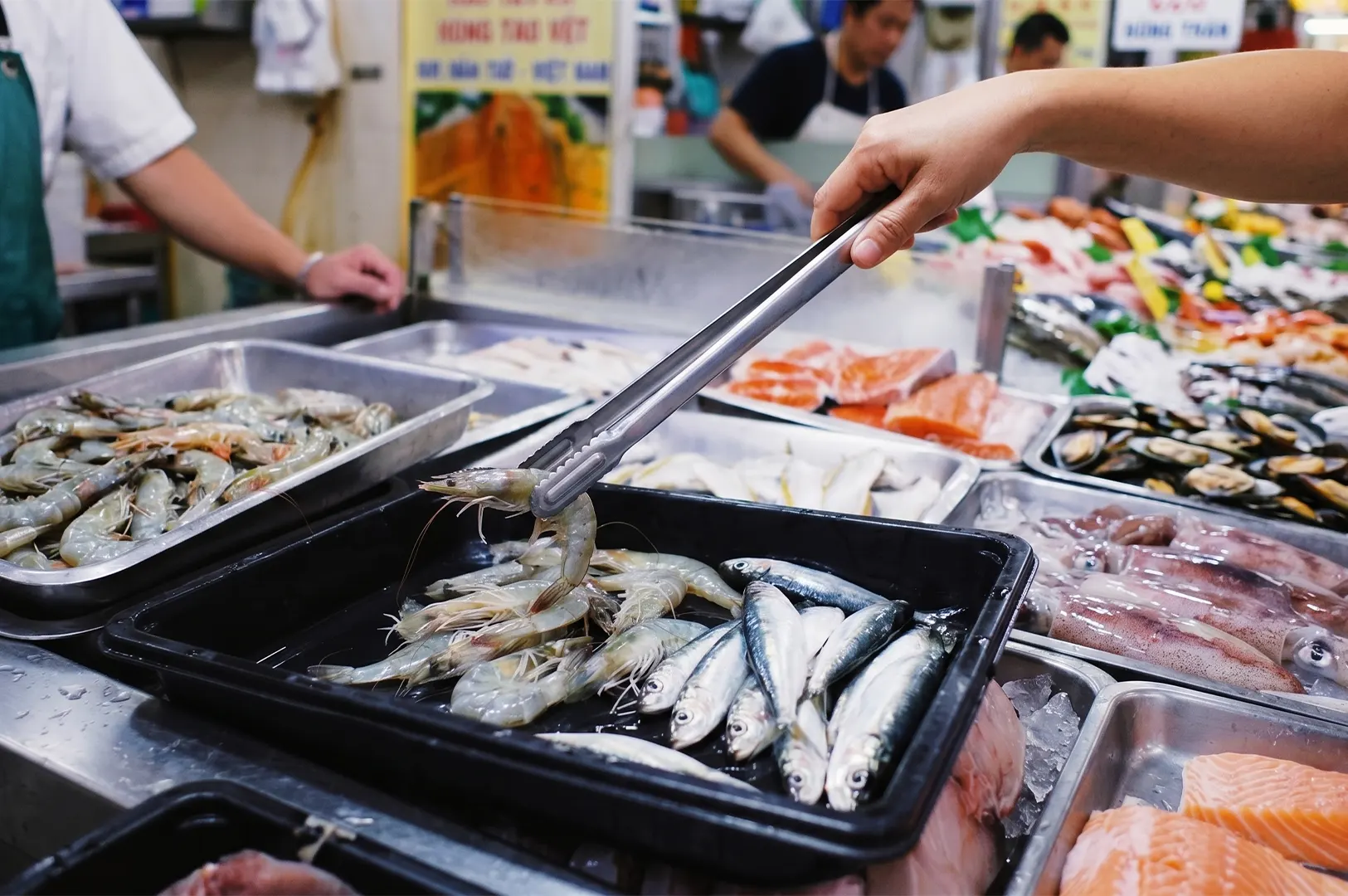 A customer using metal tongs to pick fresh prawns from a tray at a seafood market stall.