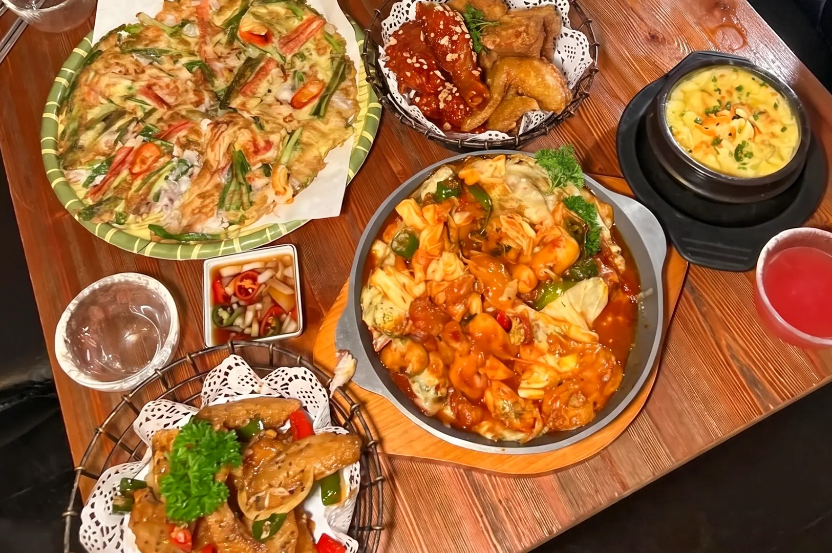 A spread of Korean dishes on a wooden table, including seafood pancake, fried chicken, spicy stir-fried meat, and steamed egg.