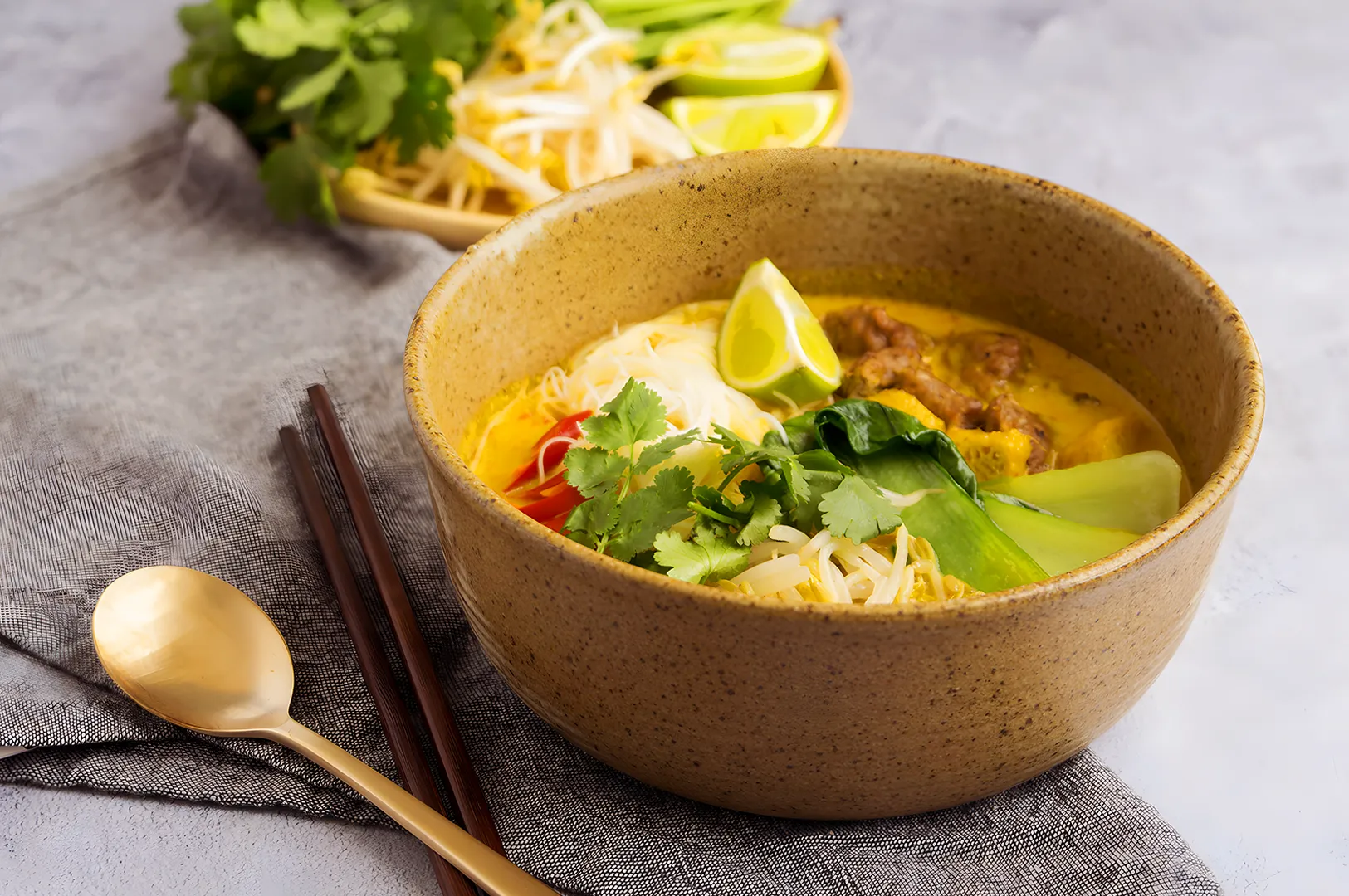 A bowl of vibrant yellow curry noodle soup with beef, topped with fresh cilantro, lime wedges, and bok choy. A golden spoon and chopsticks rest beside it.
