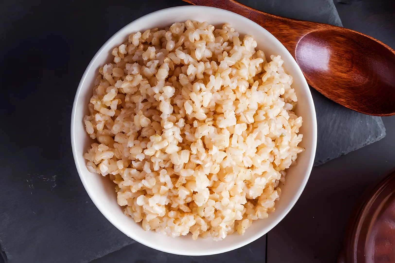 A bowl of fluffy cooked brown rice sits on a dark slate surface, accompanied by a wooden spoon. The rice is light brown and appears warm and inviting.