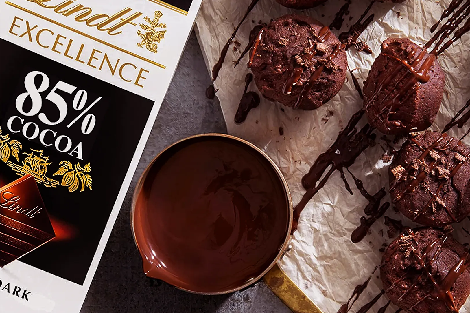 Chocolate delight scene featuring a Lindt 85% cocoa bar, a bowl of molten chocolate, and chocolate muffins drizzled with chocolate on parchment. Luxurious and rich.