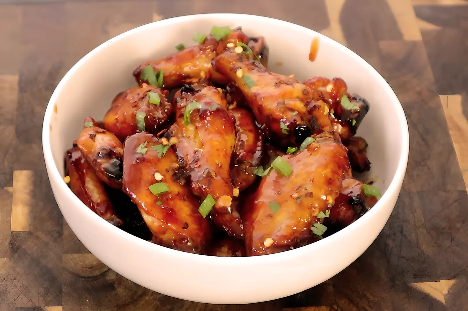 A bowl of glazed chicken wings garnished with chopped green onions. The wings are golden and caramelized, conveying a savory, appetizing appearance.