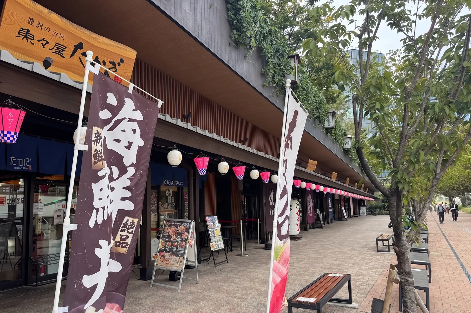 A row of traditional restaurants with banners and paper lanterns along a pedestrian walkway.