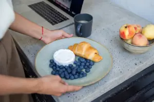 A person placing a plate with a croissant, blueberries, and brie cheese on a desk next to a laptop, coffee mug, and a bowl of peaches.