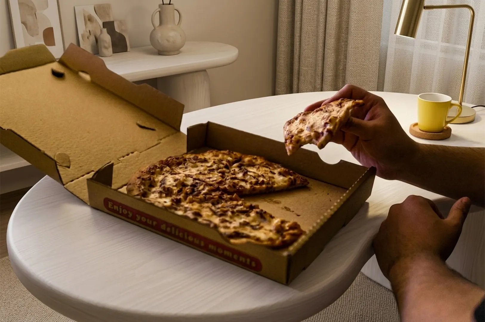 A person holding a slice of pizza over an open cardboard box on a white table in a cozy room.