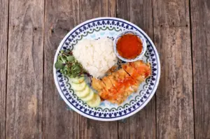 A crispy fried chicken cutlet served with heart-shaped white rice, sliced cucumbers, and sweet chili sauce on a decorative plate over a rustic wooden background.