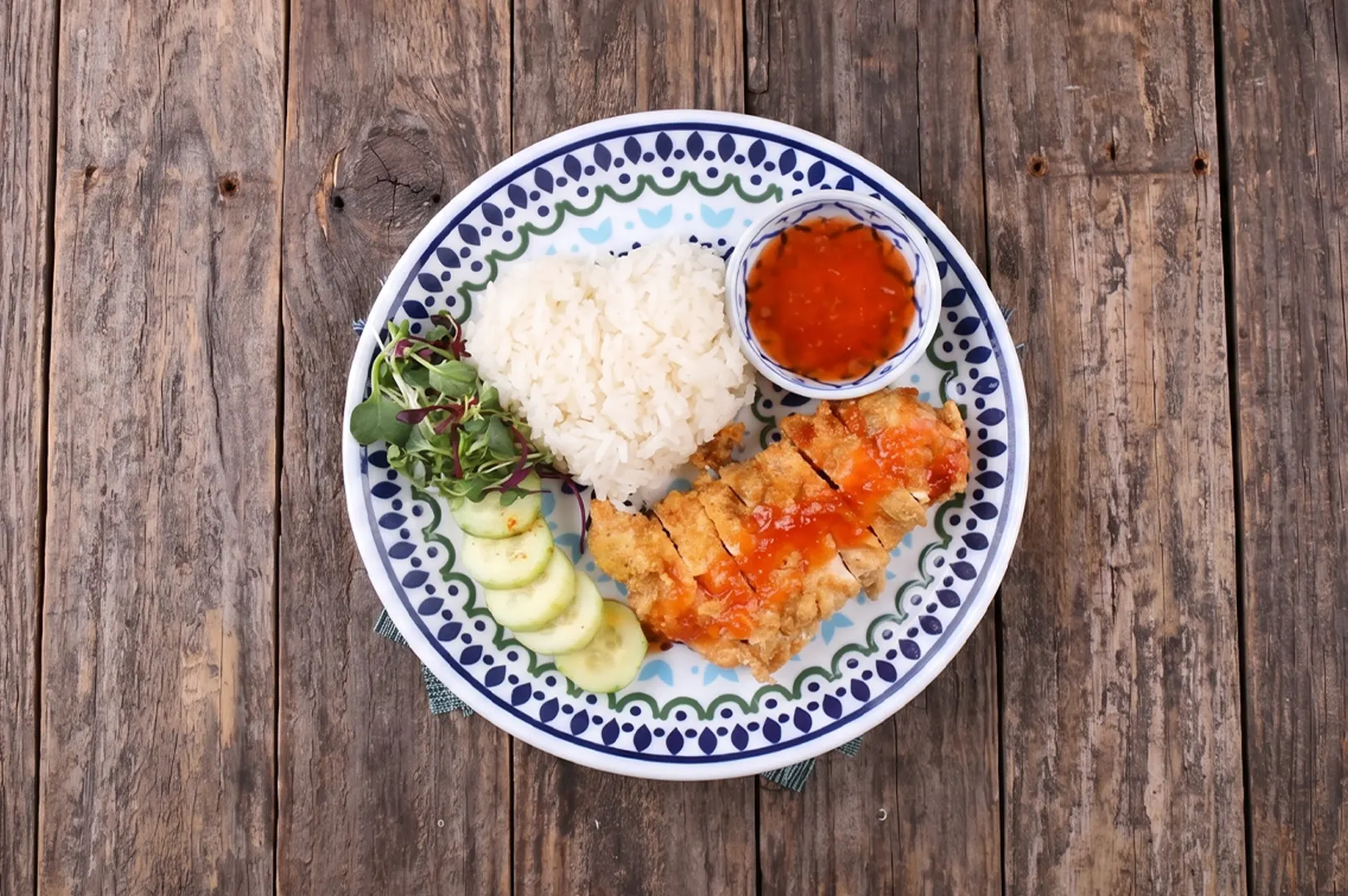 A crispy fried chicken cutlet served with heart-shaped white rice, sliced cucumbers, and sweet chili sauce on a decorative plate over a rustic wooden background.