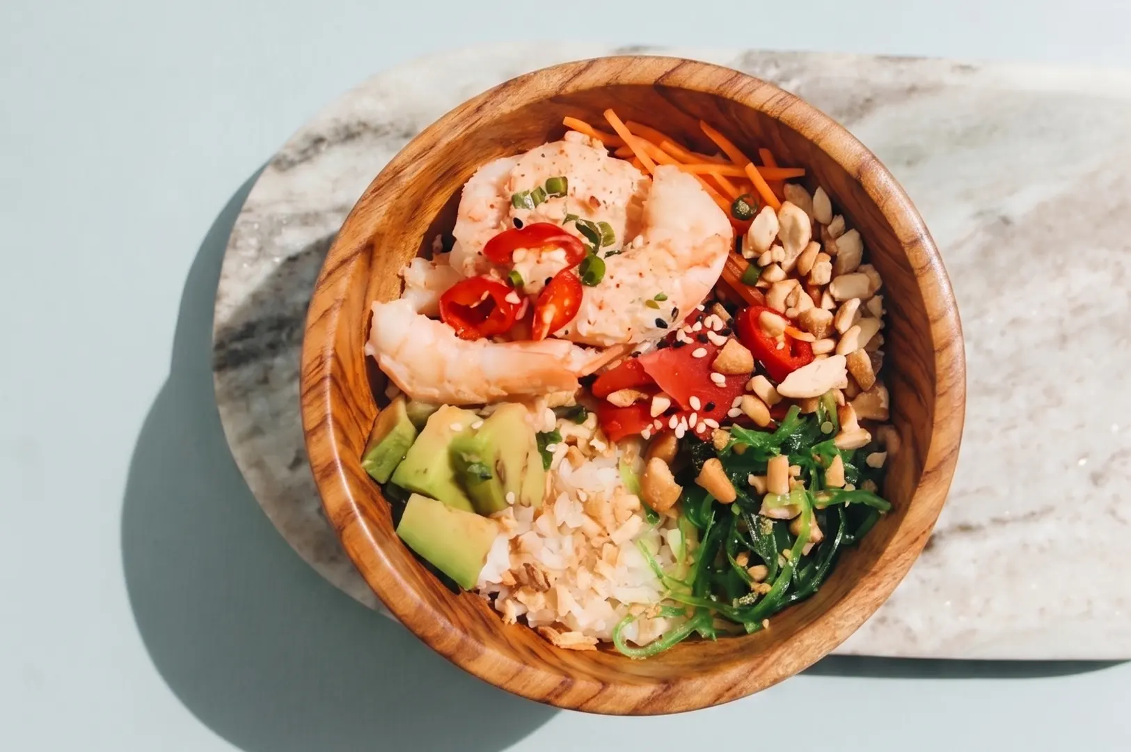 Fresh shrimp poke bowl with avocado, seaweed salad, carrots, and peanuts in a wooden bowl.