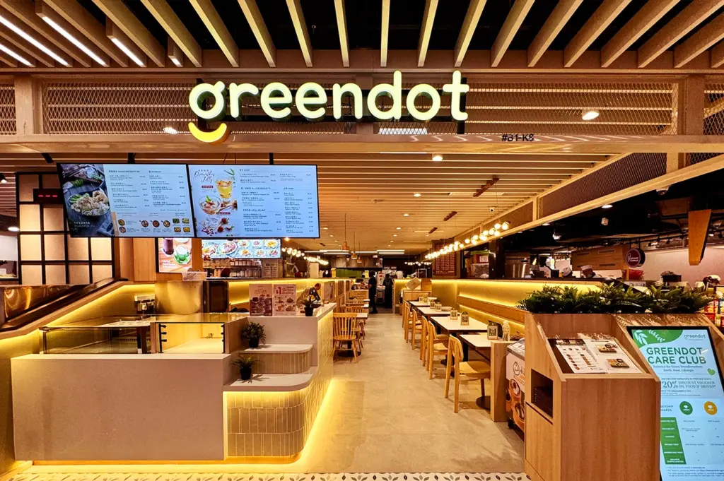 Modern restaurant interior with a sign reading "greendot" in soft yellow lighting. Wooden furnishings and digital menu boards create a warm, welcoming atmosphere.
