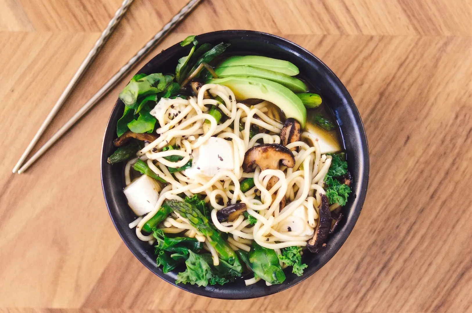 Vegetable ramen bowl with tofu, avocado, shiitake mushrooms, and kale on a wooden table.