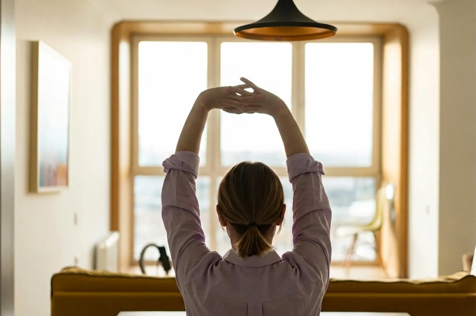 Rear view of a woman stretching her arms upward in a bright, modern living room with large windows.