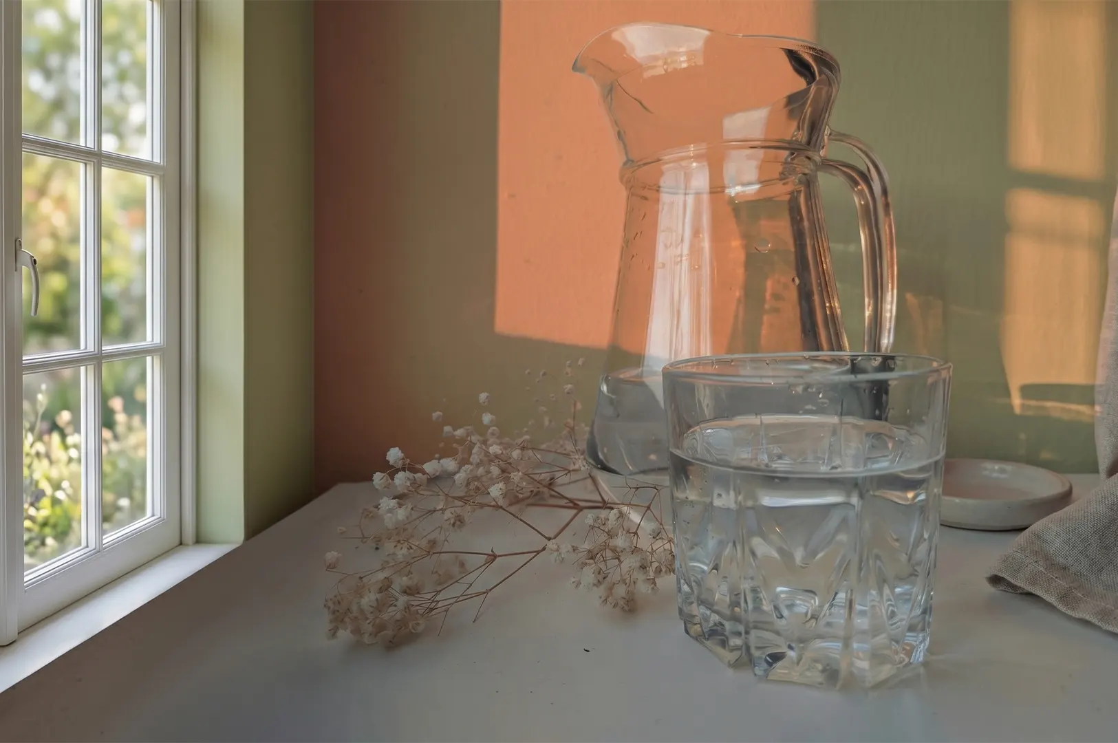 Glass pitcher and a glass of water on a white table next to a window with warm afternoon sunlight and dried baby's breath flowers.