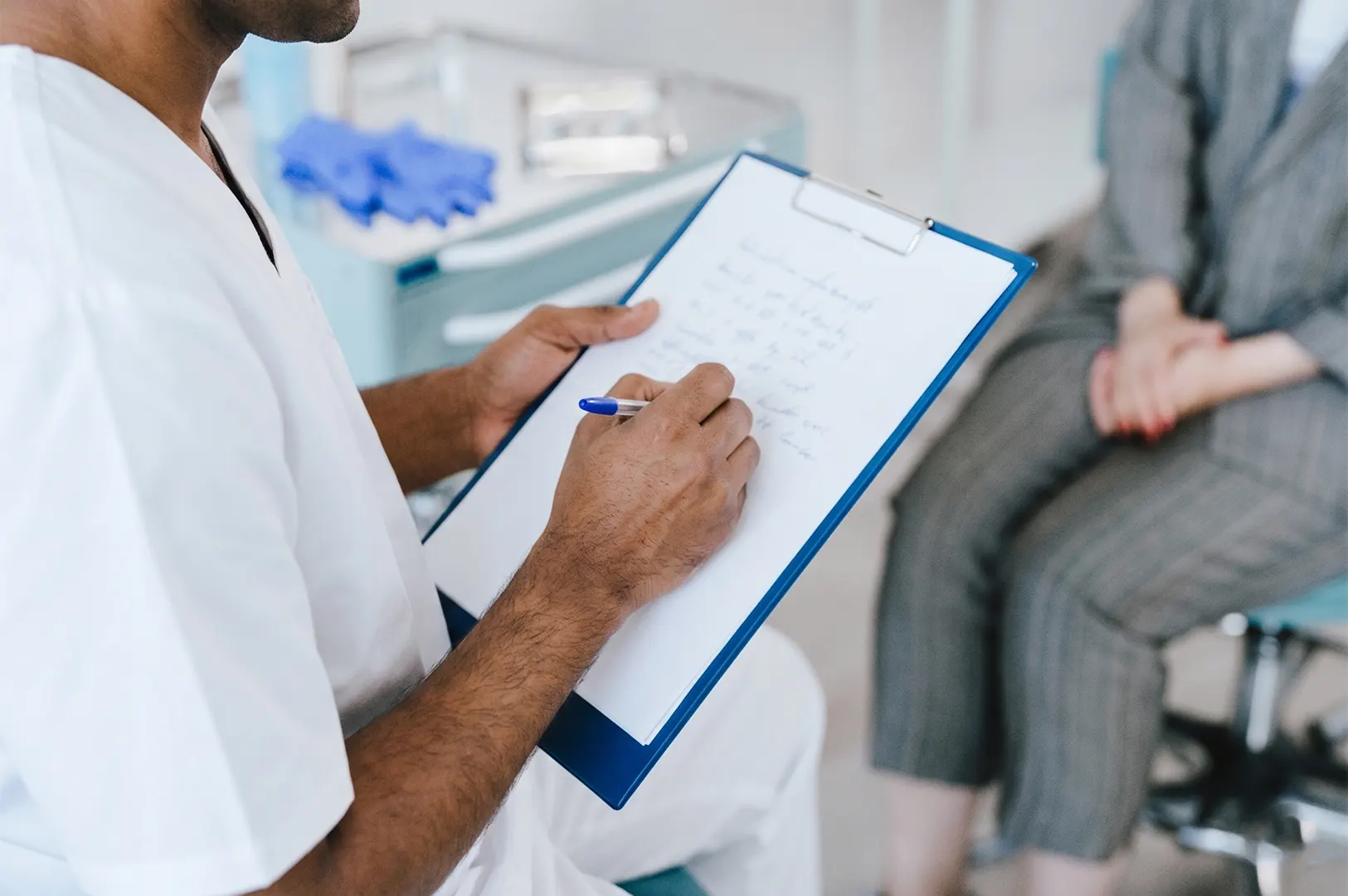 Healthcare professional in white scrubs taking medical notes on a clipboard during a patient consultation.