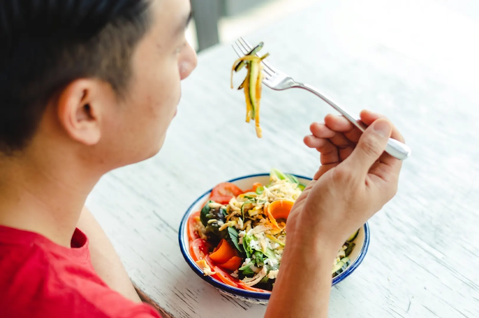 A man eating a healthy vegetable salad with zucchini noodles and tomatoes from a bowl at a white table.
