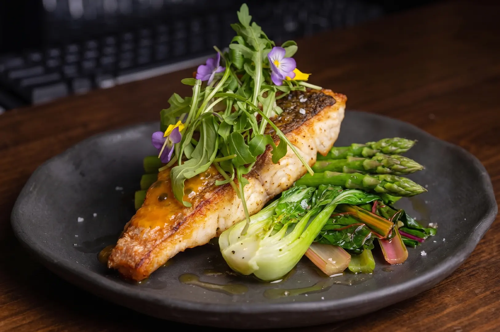 Pan-seared fish fillet served with asparagus, bok choy, and Swiss chard, topped with fresh arugula and edible purple flowers on a dark plate.