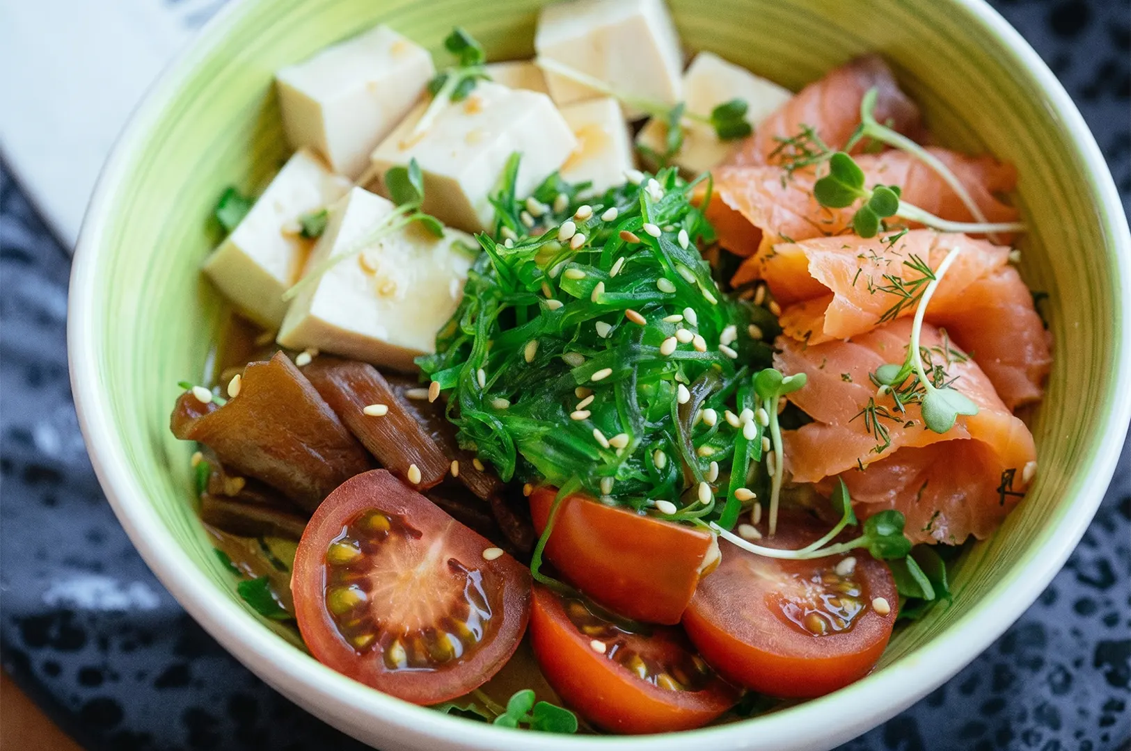 A poke bowl featuring smoked salmon, cubed tofu, seaweed salad topped with sesame seeds, and sliced cherry tomatoes in a light green bowl.
