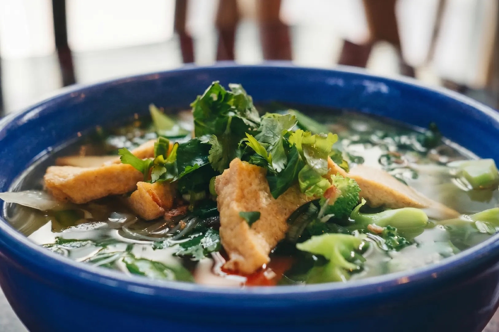 A blue bowl of tofu pho noodle soup topped with fresh cilantro and broccoli in a clear broth.