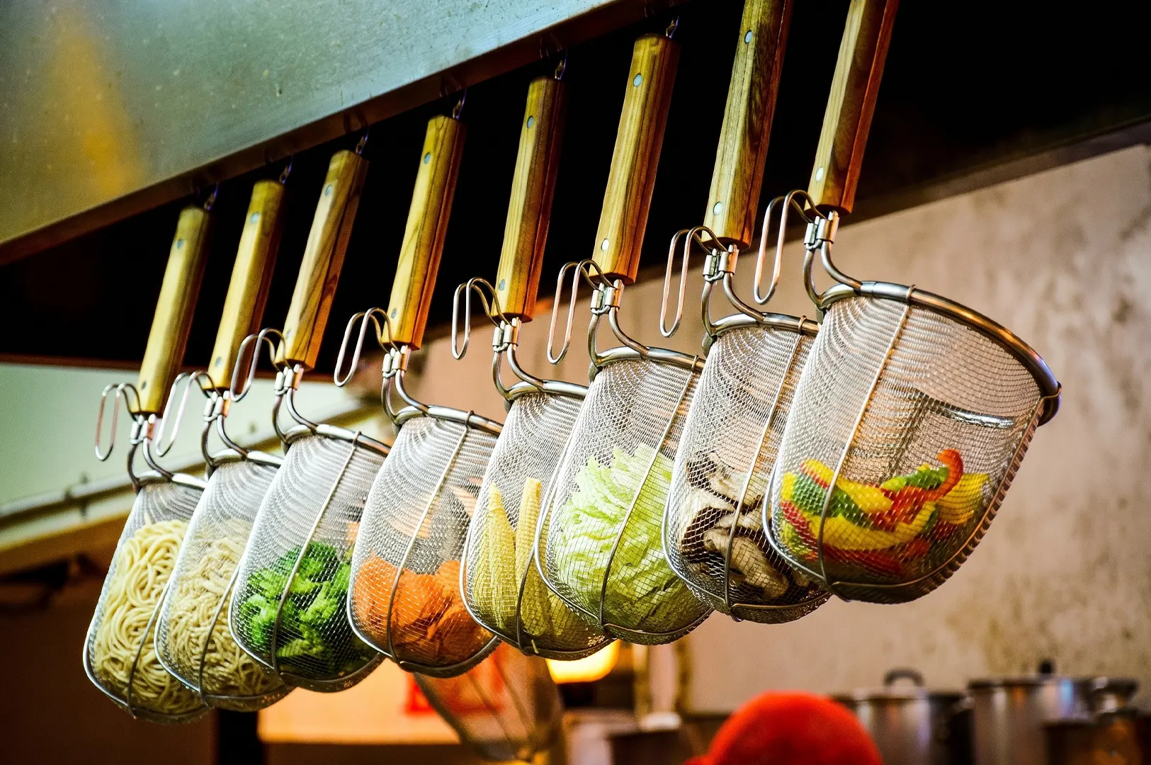 A row of stainless steel noodle strainers with wooden handles, filled with fresh ramen noodles, broccoli, carrots, corn, cabbage, and mushrooms.