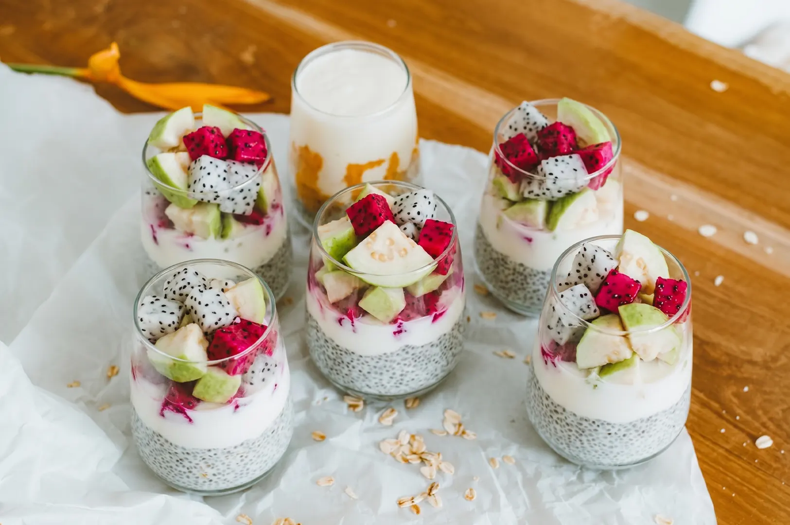 Five glasses of layered chia seed pudding topped with white yogurt and fresh cubes of red and white dragon fruit and guava.