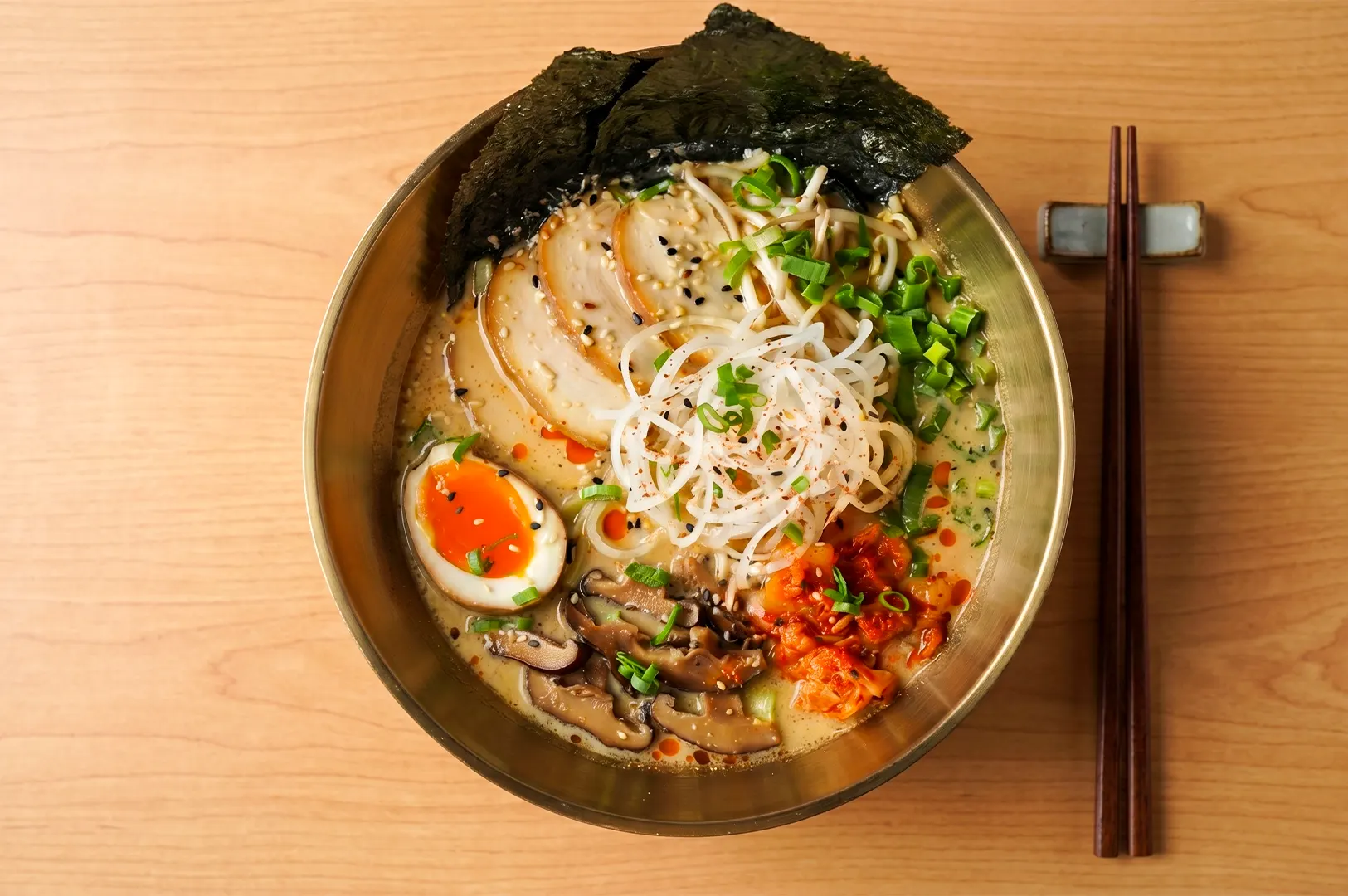 Top-down view of a ramen bowl with chashu pork, a soft-boiled egg, nori, kimchi, and shiitake mushrooms in a golden bowl.