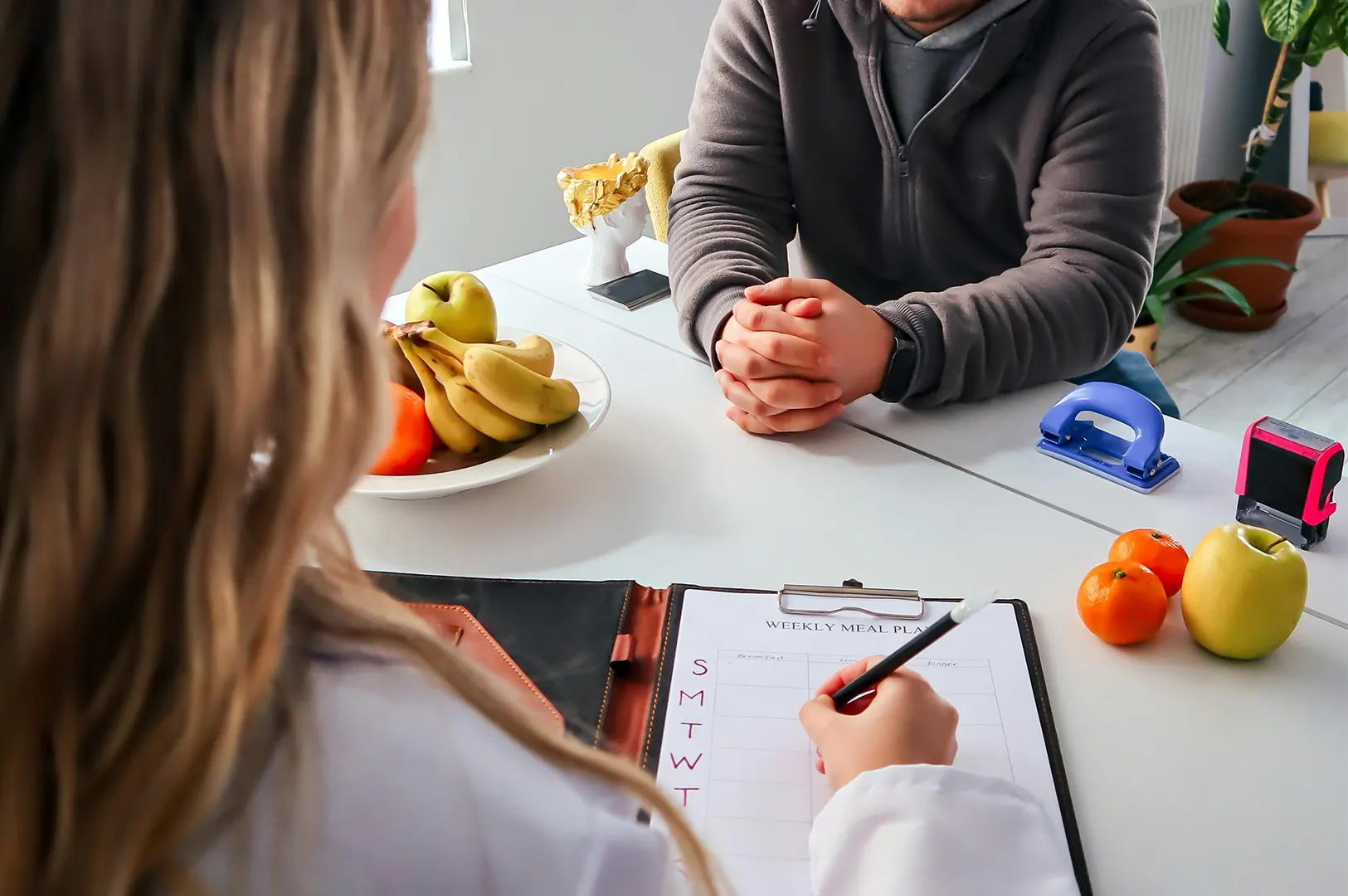 A nutritionist or dietitian filling out a weekly meal plan during a consultation with a client, with fresh fruit like apples and bananas on the table to promote healthy eating habits.