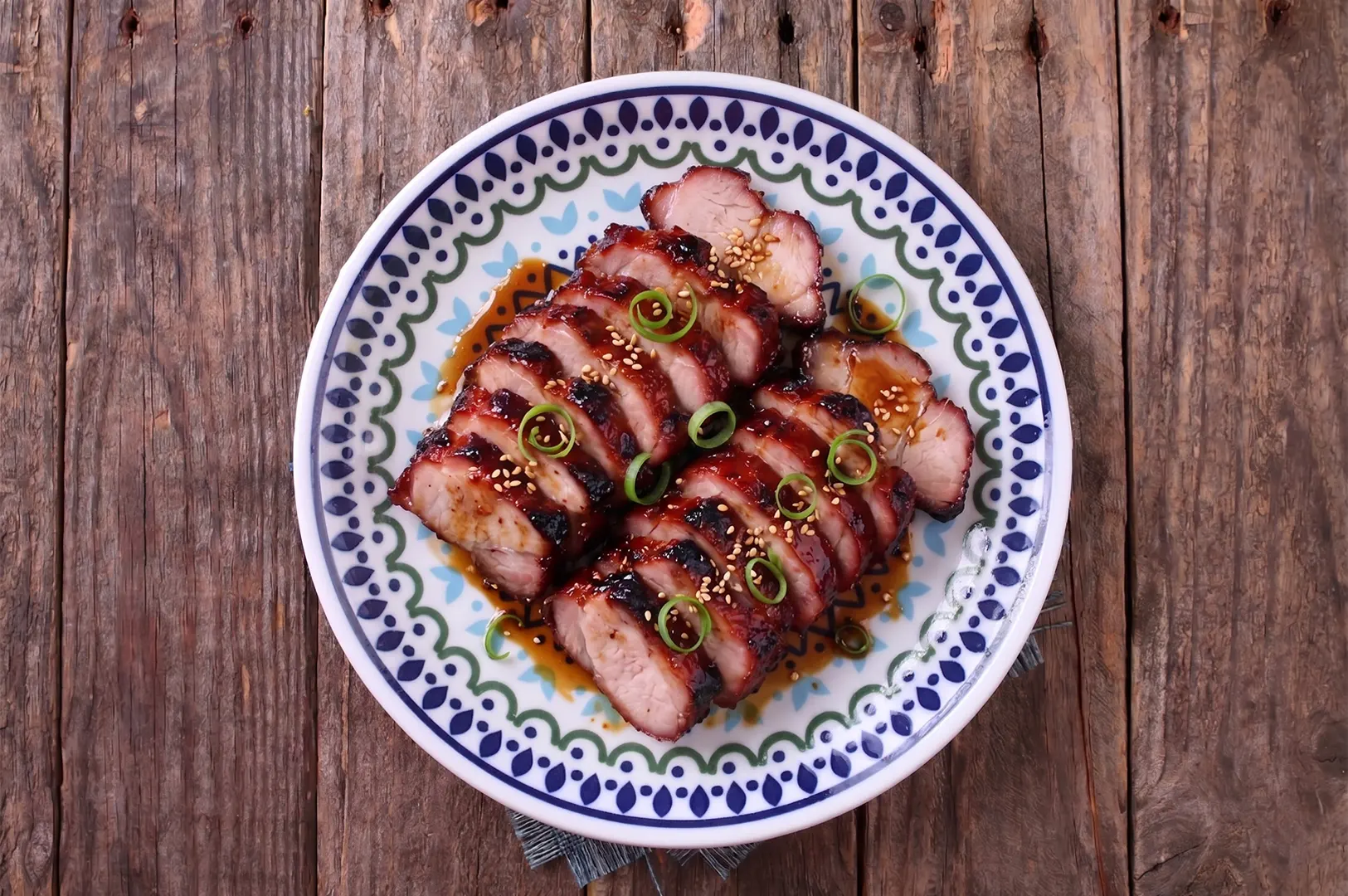 Top-down view of sliced Chinese BBQ pork (char siew) with a glossy glaze, garnished with sesame seeds and green onions on a patterned plate over a rustic wooden table.