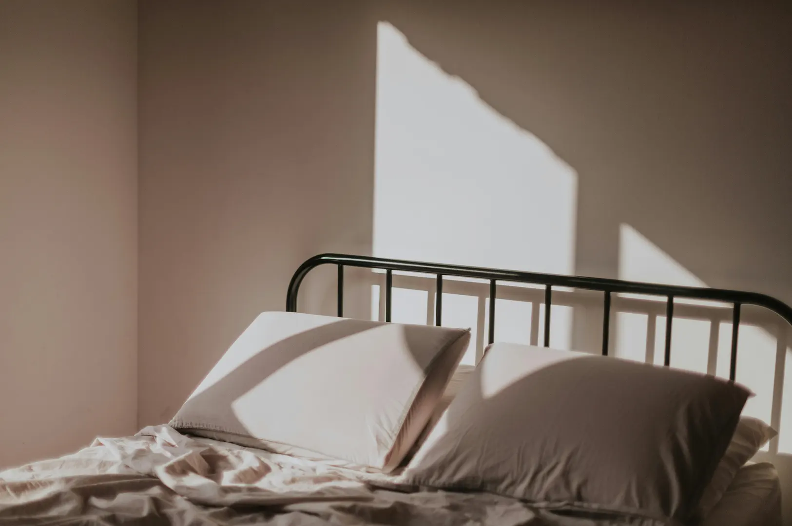 Minimalist bedroom interior featuring a black metal bed frame with white pillows and sunlight casting shadows on the wall.