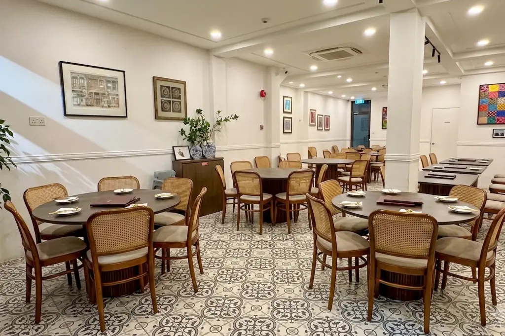 Bright interior dining room of Chilli Padi Nonya Restaurant featuring round wooden tables, cane back chairs, and traditional patterned floor tiles.