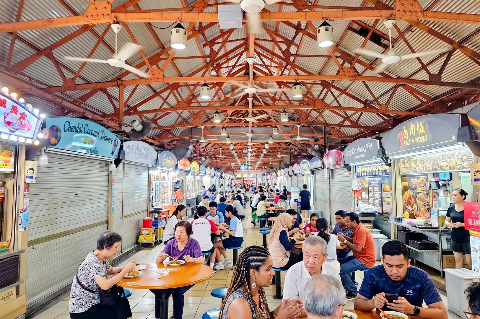 Diverse group of people eating at tables in a bustling indoor hawker center.