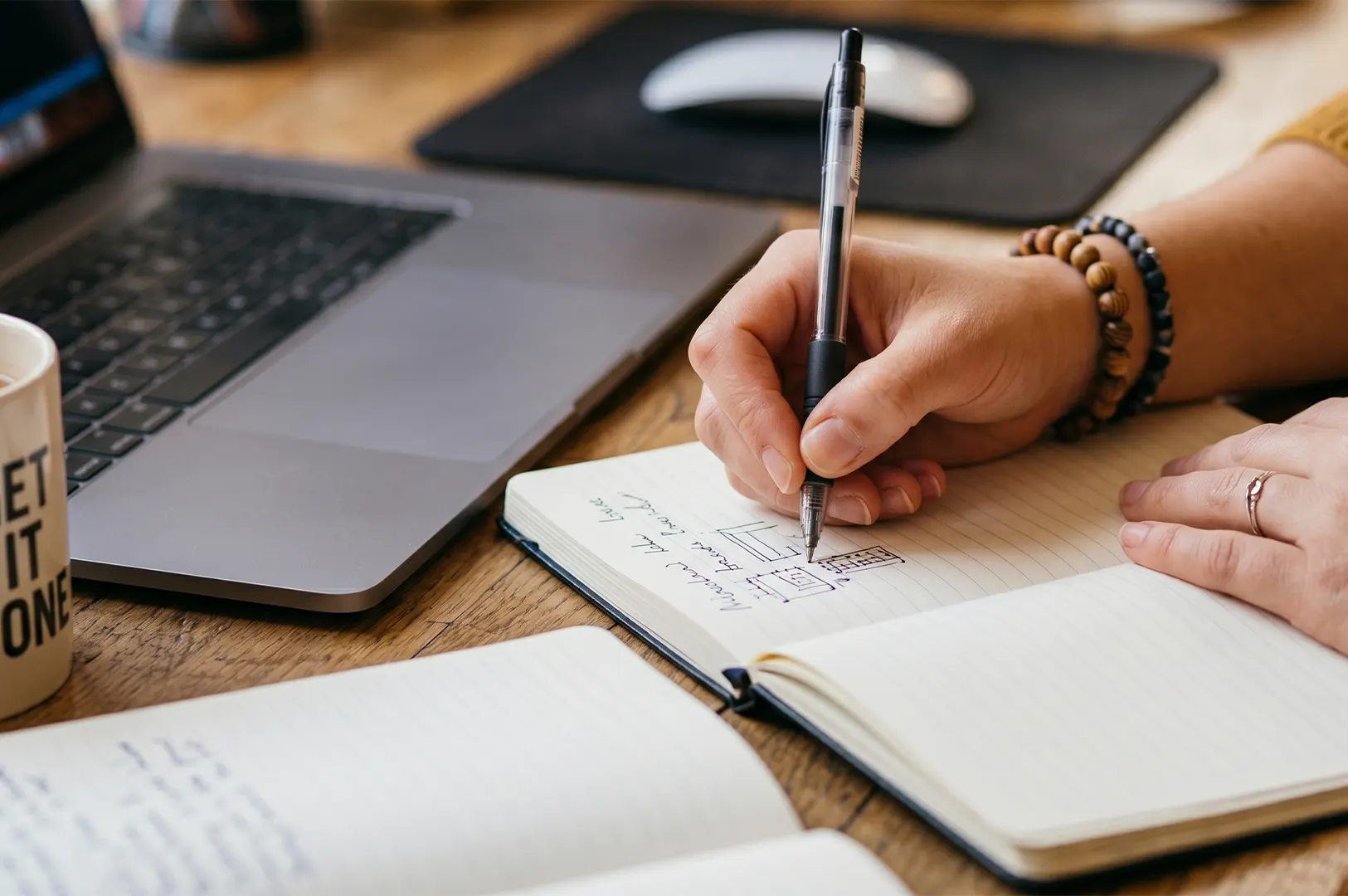 A person writing notes and sketching diagrams in a notebook next to an open laptop on a wooden desk.