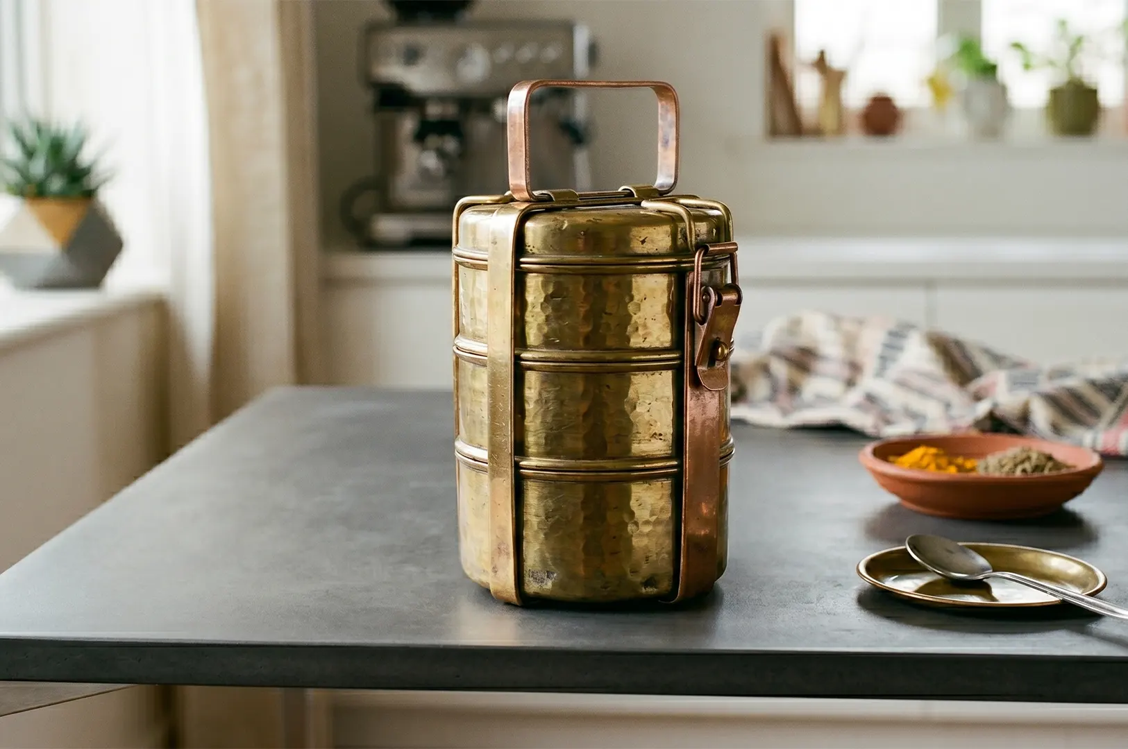 Traditional brass 3-tier tiffin carrier on a dark wooden table in a bright kitchen.